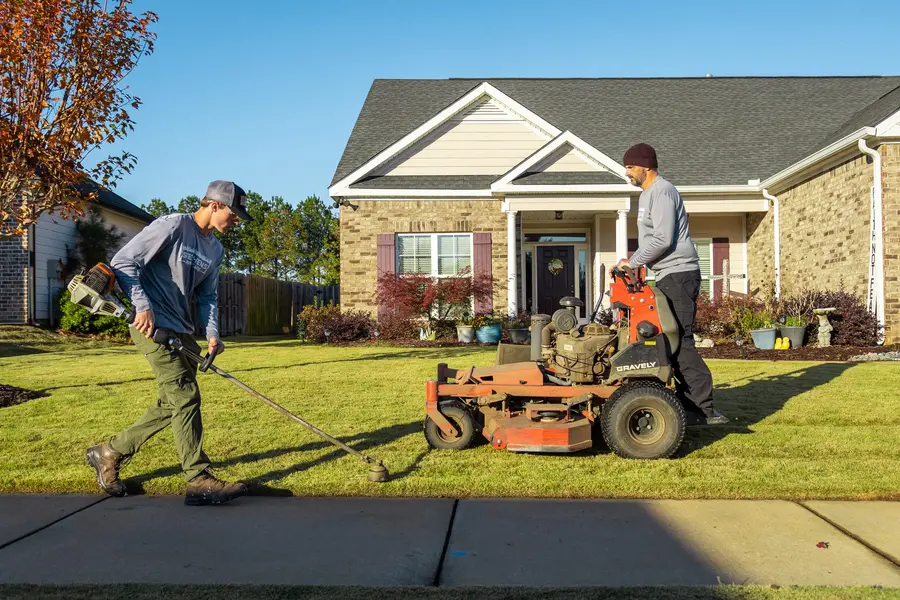 Two Stephens Lawn Care crew members trimming and mowing the same front lawn in low evening light